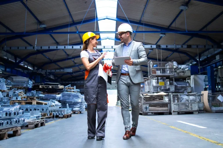 supervisor-gerente-e-trabalhador-industrial-uniformizado-caminhando-no-grande-salao-de-uma-fabrica-de-metal-e-falando-sobre-o-aumento-da-producao
