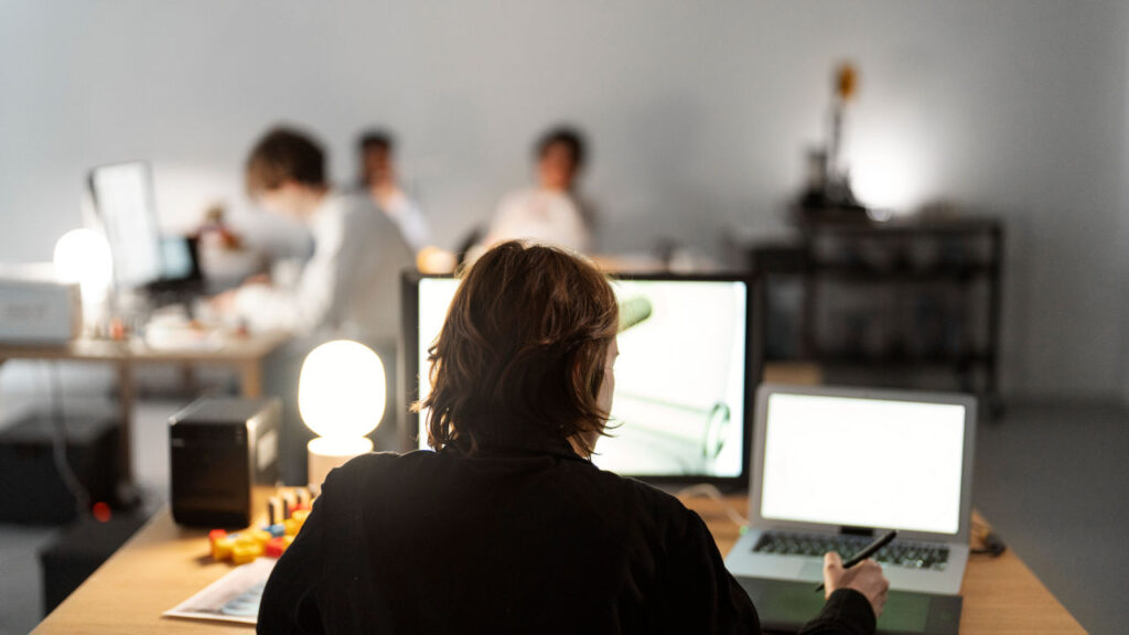 Pessoa utilizando dois monitores e um laptop em um ambiente de escritório colaborativo.