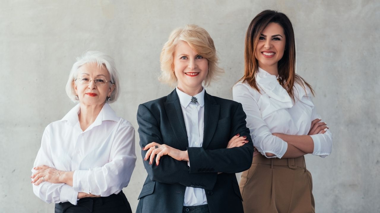 três mulheres de diferentes idades, sorridentes e de braços cruzados.